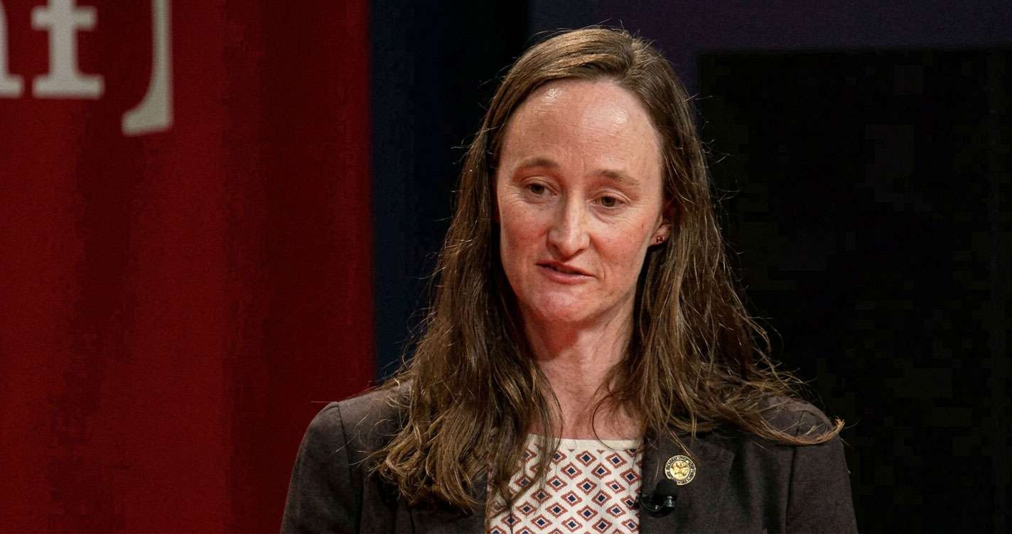 Woman with long brown hair, wearing a blazer and patterned top, speaking at a stage with a red backdrop.