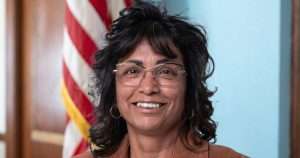 Smiling woman with dark hair and glasses, wearing a brown jacket, in front of an American flag.