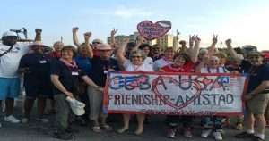 Group of diverse people at a rally holding a banner reading 'CUBA-USA Friendship-Amistad' with a heart and flags in the background.