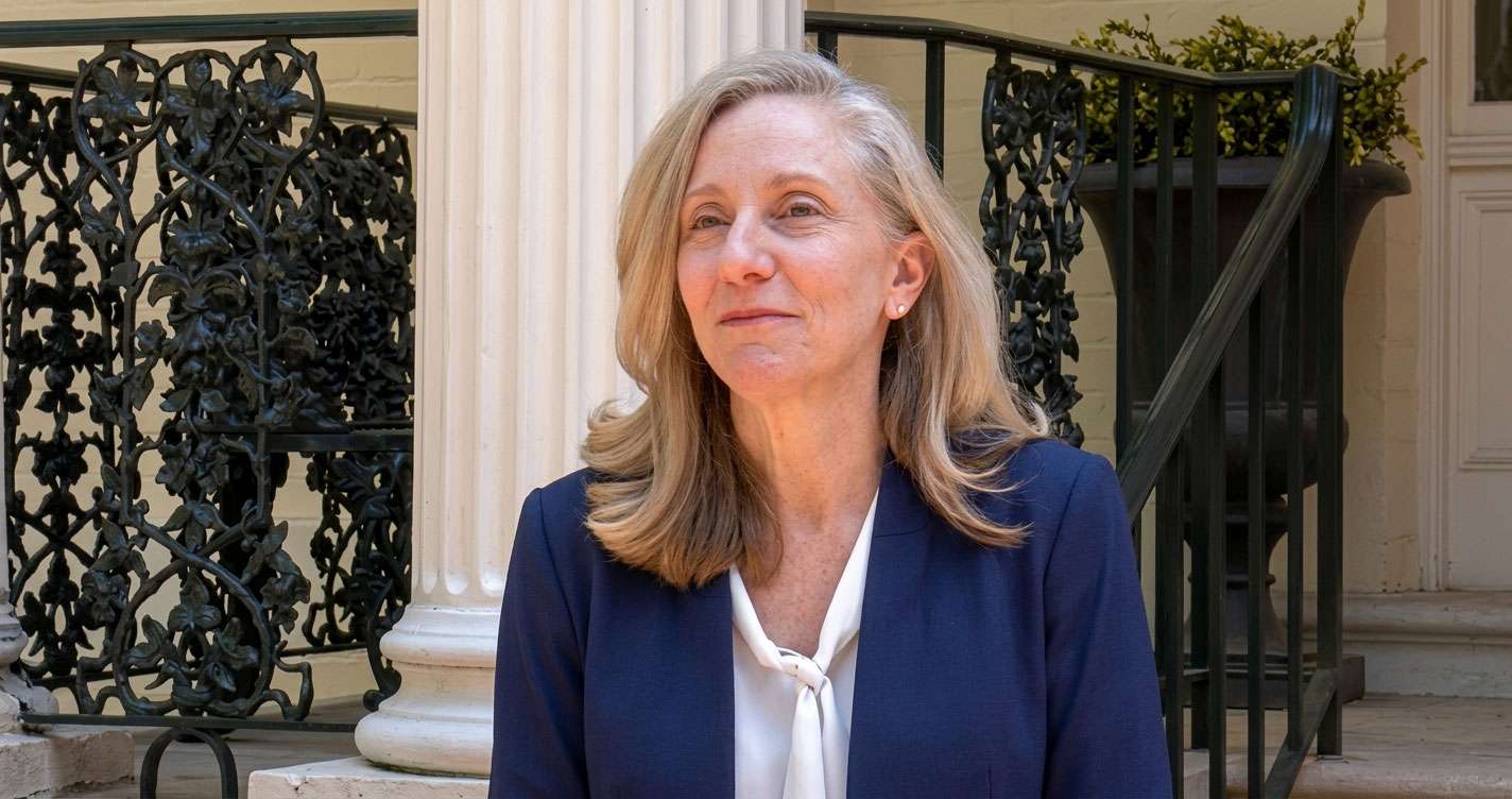 Professional woman with blonde hair, wearing a navy blazer and white blouse tied at the neck, standing outside a building with decorative iron railing and a stone column.