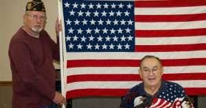 Two men pose in front of a large American flag; a standing veteran in a burgundy sweater and cap beside the pole, and a seated man in a star‑spangled shirt.