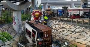 Overturned brown bus wedged in a rocky ditch beneath a highway, with rescue workers and tow trucks on scene.