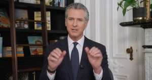 Professional man in a dark suit and tie speaks to the camera, gesturing with his hands in a library-like room with bookshelves behind him.