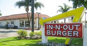 In-N-Out Burger sign with a large yellow arrow, set against a sunny fast-food building and palm trees in the background, exterior view of the restaurant.