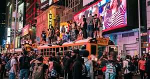 Crowded city street at night with a yellow school bus covered in people on its roof and many pedestrians on the ground beneath, illuminated by large billboards.