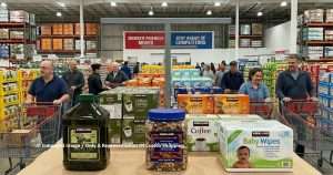 Shoppers push grocery carts through a busy warehouse-style store aisle with bulk coffee, oil, and snacks on display in the foreground.