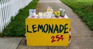Bright yellow lemonade stand on a sidewalk with cups, a pitcher of lemonade, lemons, and a jar of money; sign reads 'LEMONADE 25¢'.