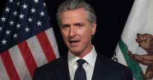 Man in a dark suit and tie speaking, with US and California flags in the background.