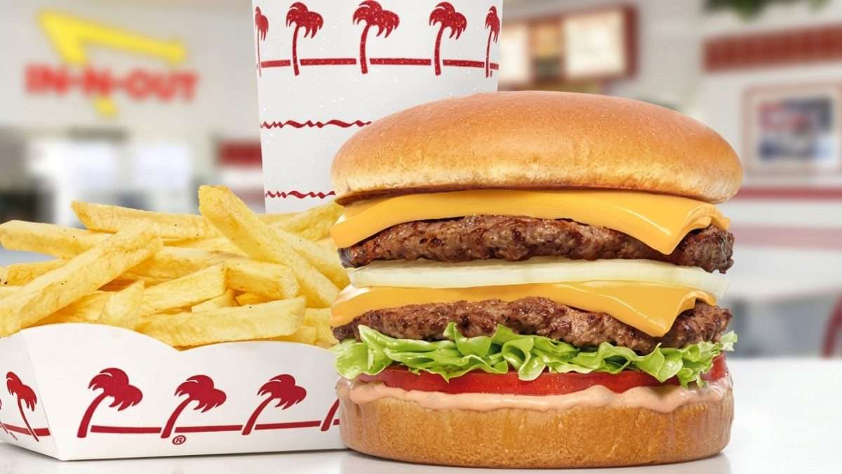 Two-patty cheeseburger with lettuce, tomato, and sauce, beside a box of fries and a drink.