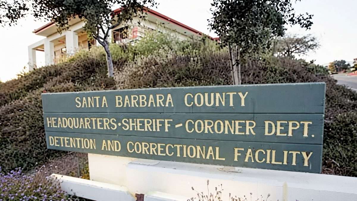 Sign reading 'Santa Barbara County Headquarters Sheriff-Coroner Dept. Detention and Correctional Facility' with shrubs and a building visible behind.